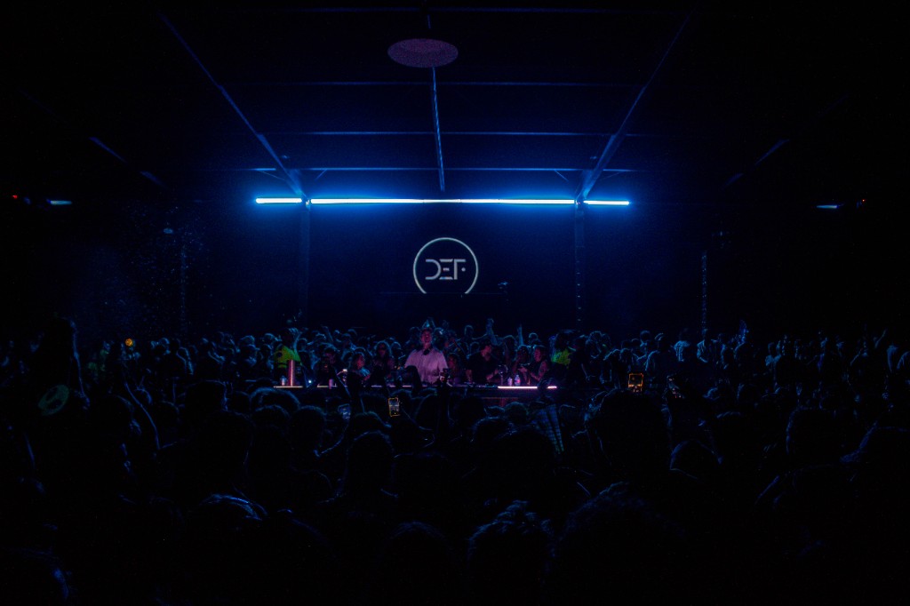 Blue neon over the warehouse, DEF logo, crowd in silhouette